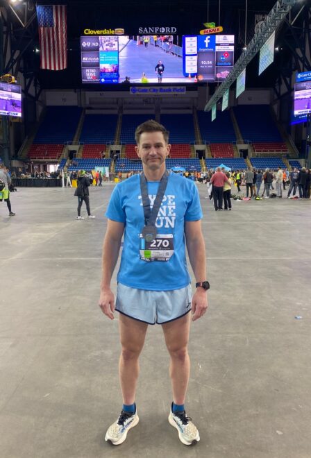 Cody stands in a stadium after a marathon, wearing his race medal and a blue Type One Run tshirt 