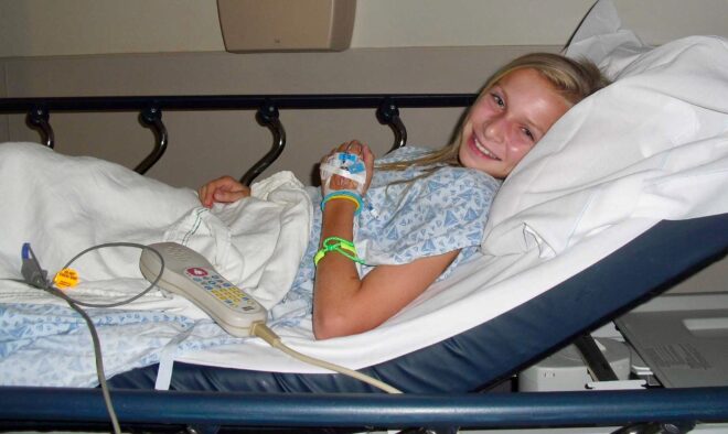 An eleven year old girl wearing a hospital gown lays in a hospital bed, an IV in her arm, but she is smiling