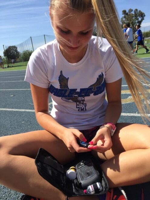 Close up photo of a teenage girl with a blonde pony tail, sitting cross legged on the ground, checking her blood sugar by pricking her finger