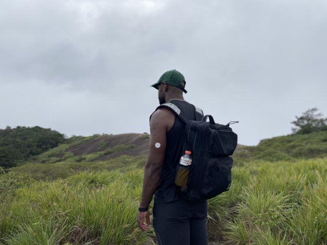Dex, wearing a backpack and continuous glucose monitor, stands in a field looking away from the camera.