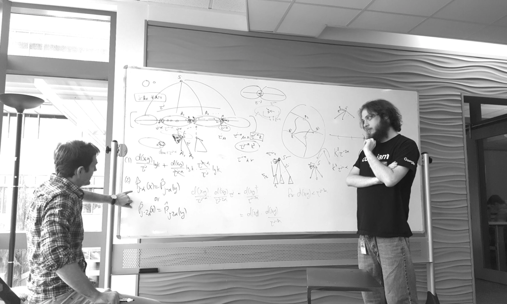 Black and white photo showing Mikey working with a colleague near a white board covered with equations